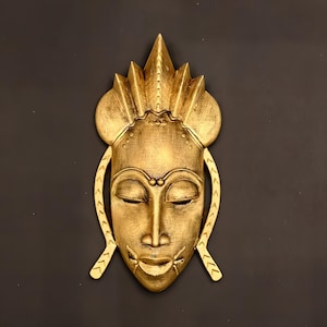 May include: A gold-colored mask with a crown-like headpiece and intricate details. The mask has a closed-eye expression and a small bead on the forehead.
