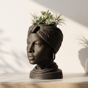 May include: A matte black head-shaped planter featuring a sculpted face wearing a headwrap, hoop earrings, and a beaded necklace. The base is decorated with a geometric chevron pattern. A collection of green succulents is planted in the top.
