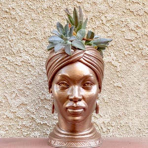 May include: A bronze-colored head-shaped planter with a turban, holding a succulent plant. The planter has detailed facial features and hoop earrings. The succulent has green and red leaves. The planter sits on a red surface against a textured wall.