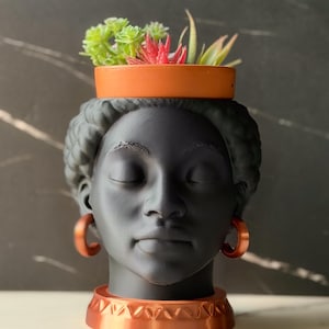 May include: A matte black head-shaped planter featuring a person with closed eyes, wearing copper-colored hoop earrings and a matching copper-colored base. The top of the head serves as a container holding several small green and red succulent plants.