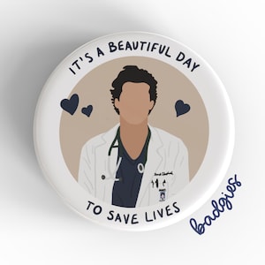 May include: A white button with a brown background featuring a cartoon illustration of a doctor in a white coat and blue scrubs. The text "It's a beautiful day to save lives" is written in black around the image. Two black hearts are on either side of the doctor. The text "badgrrls" is written in blue at the bottom right of the button.