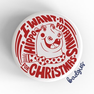 May include: A white button with red text that reads "I want a hippopotamus for Christmas" with a cartoon hippopotamus wearing a red and white scarf. The text "badges" is in blue at the bottom.