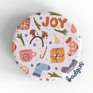 May include: A white round button with a Christmas themed design. The button features a variety of festive elements, including presents, a rocking horse, a cup of hot cocoa, candy canes, and a Christmas tree. The word "JOY" is written in large letters at the top of the button. The text "badgriss" is written in blue at the bottom of the button.