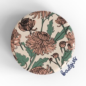 May include: A round phone grip with a floral pattern. The pattern features pink and green chrysanthemums on a cream background. The text "badgirls" is written in blue cursive on the bottom right corner of the grip.