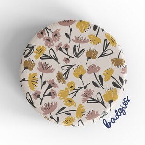 May include: A round button with a floral pattern. The button features a white background with yellow and pink flowers and black stems. The text "badgriss" is written in blue at the bottom of the button.
