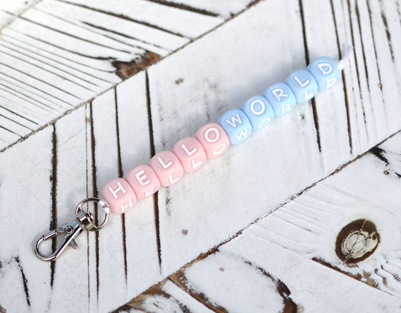 Pu&ograve; includere: Portachiavi in silicone rosa e blu con le parole "HELLO WORLD" scritte. Il portachiavi ha una chiusura in metallo argentato.