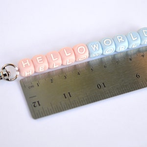 Pu&ograve; includere: Un portachiavi con perline rosa e blu con le parole "HELLO WORLD" scritte. Il portachiavi &egrave; misurato con un righello, mostrando che &egrave; lungo circa 20 cm.