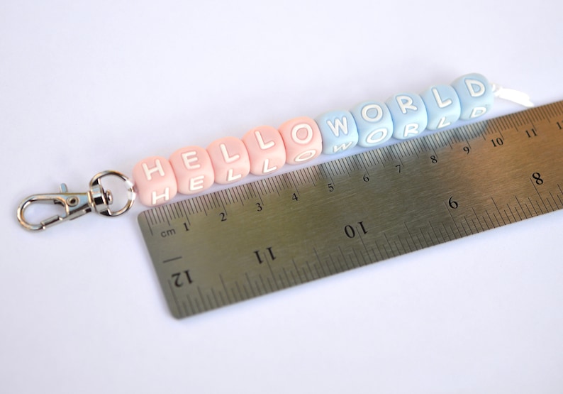 Pu&ograve; includere: Un portachiavi con perline rosa e blu con le parole "HELLO WORLD" scritte. Il portachiavi &egrave; misurato con un righello, mostrando che &egrave; lungo circa 20 cm.
