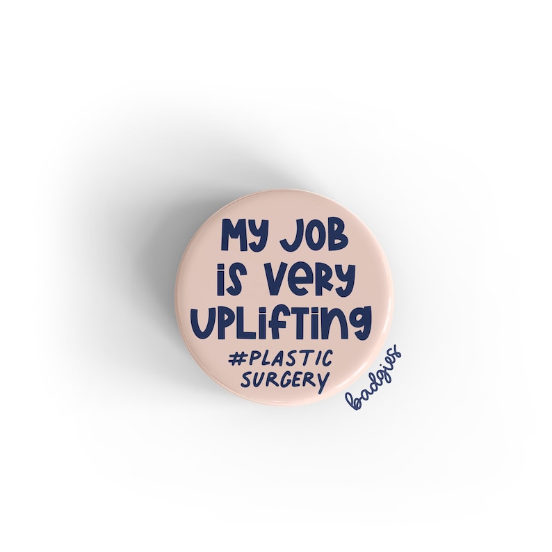 K&ouml;nnte beinhalten: Ein rosa Knopf mit dem Text "My job is very uplifting #plastic surgery" in dunkelblauer Schrift.