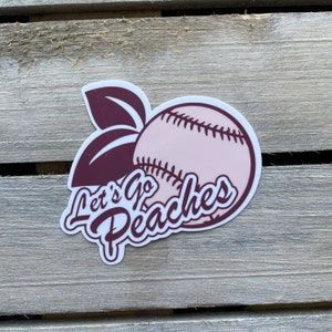 May include: A pink peach with a baseball stitched pattern and a maroon leaf. The peach has the text "Let's Go Peaches" in maroon cursive.