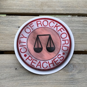 May include: A white sticker with a red border and the text "City of Rockford Peaches" in a red circle. The circle also features a black scale design in the center.