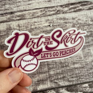 May include: A white sticker with maroon text that reads "Dirt in the Skirt" with a baseball and a leaf. The text below the baseball reads "Let's Go Peaches".