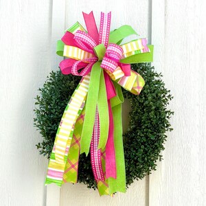 May include: A green boxwood wreath adorned with a large bow. The bow features ribbons in bright pink, lime green, and yellow with striped and plaid patterns. The wreath is hanging on a white door.