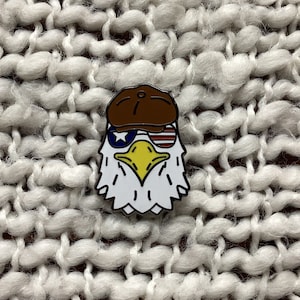 May include: A white, yellow, and brown enamel pin featuring an eagle wearing sunglasses and a brown cap. The sunglasses have a red and white striped design. The eagle has a blue and white star design on its chest.