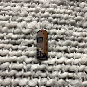 May include: A brown and orange enamel pin with a cartoon illustration of a room with a lamp, a window, and the text "PARASOL 22:47".