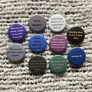 May include: Ten round button pins with various colors and text. The text on the pins includes quotes from the TV show The Office. The quotes include: "Save Bandit!" "Bears. Beets. Battlestar Galactica." "There's a lot of beauty in ordinary things. Isn't that kind of the point?" "Identity theft is not a joke, Jim!" "The people person's paper people." "And you, my friend, would be da belle of da ball!" "Limitless paper in a paperless world." "I hate so much about the things that you choose to be." "My whole life is a booger bubble." "Scranton, PA"