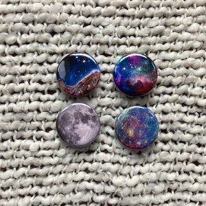 May include: Four round buttons with different space-themed images. The buttons are on a white knitted surface. The images include a nebula, a moon, and two different galaxies.