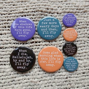 May include: A collection of round pinback buttons in various colors, each with a different phrase in white text. The phrases include "When the shadows of this life have gone, I'll fly away" and "When I die, Hallelujah, by and by, I'll fly away."