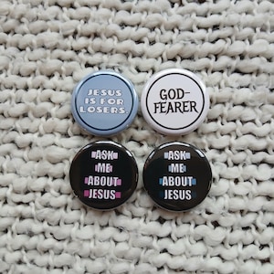 May include: Four round buttons with text. One light blue button reads "JESUS IS FOR LOSERS." A white button reads "GOD-FEARER." Two black buttons say "ASK ME ABOUT JESUS" in pink and blue.