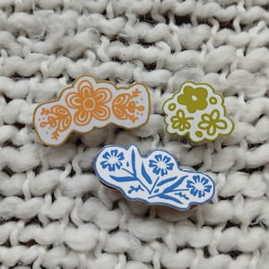 May include: Three enamel pins with floral designs. One pin is orange and white, another is green and white, and the third is blue and white. The pins are on a white, textured surface.