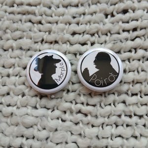 May include: Two white circular pinback buttons with black silhouettes of literary characters. One button features a woman's profile with the name "Marple" in a curved font. The other button shows a man's profile with the name "Poirot".