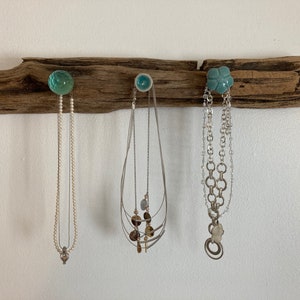May include: A driftwood jewelry holder with three necklaces hanging from it. The holder has three hooks, each with a different colored glass or ceramic knob. The necklaces are silver and have different styles.