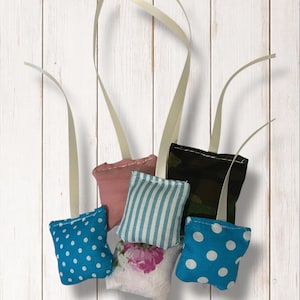 Può includere: Una collezione di piccoli sacchetti decorativi in tessuto con vari motivi e colori, tra cui pois blu, rosa, floreale, a righe e mimetico. Ogni sacchetto ha un nastro color crema per appenderlo.