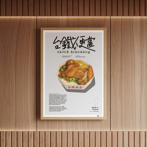 Puede incluir: Un póster enmarcado con una ilustración de una caja bento taiwanesa. El póster tiene caracteres chinos y las palabras "táitiě biàndāng". La ilustración de la caja bento muestra arroz, pollo frito, verduras y un huevo cocido. El póster está en una pared de madera.