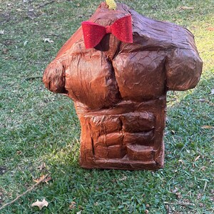 May include: A brown, sculpted torso with a red glitter bow tie. The sculpture has defined abdominal muscles and a textured surface. A small, gold-colored rope is at the top of the sculpture. The sculpture is outdoors on a grassy surface.