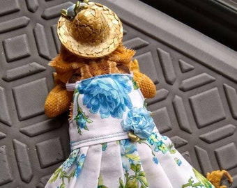 May include: A bearded dragon wearing a white dress with blue floral print and a straw hat. The dress has a blue ribbon bow on the front.