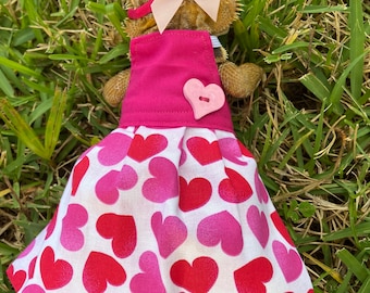 May include: A pink and white heart-patterned dress with a pink bow and a pink heart button. The dress is worn by a bearded dragon.