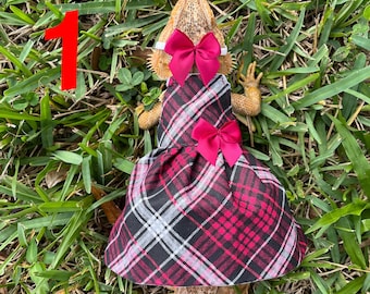 May include: A black, pink, and white plaid dress with a pink bow on the front. The dress is worn by a bearded dragon with a pink bow on its head. The dragon is sitting on green grass.