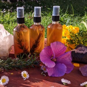 May include: Four amber glass bottles with black spray tops filled with a brown liquid and dried herbs. The bottles are arranged in a row on a wooden surface with a clear quartz crystal, a purple flower, and other crystals.