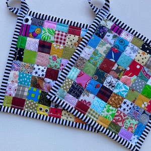 May include: Two colorful patchwork potholders with black and white striped borders and hanging loops. The potholders are made of small, square fabric pieces in various colors and patterns, including floral, stars, and polka dots.