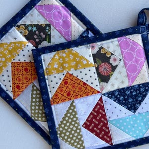 May include: Two square quilted potholders with a patchwork design. The potholders feature a variety of fabrics in colors like yellow, orange, pink, and blue, with a dark blue border and a loop for hanging.