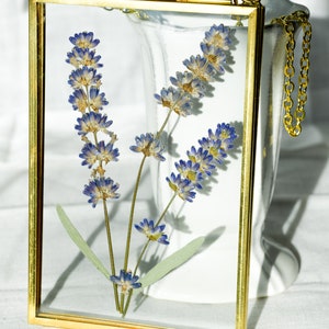 May include: A gold-framed pressed flower artwork featuring dried lavender. The lavender has blue-purple flowers and green leaves, encased in a rectangular frame. A gold chain is attached for hanging. The frame is approximately 15 cm x 20 cm.