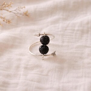 Puede incluir: Un anillo plateado con dos cuentas esféricas negras. El diseño del anillo incorpora detalles en forma de flecha, creando un accesorio único y elegante. El anillo se muestra sobre una tela texturizada de color blanco roto.