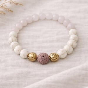 Puede incluir: Una pulsera de cuentas redondas blancas y rosa claro, con tres cuentas de acento. Las cuentas de acento incluyen dos cuentas doradas texturizadas y una cuenta malva porosa. La pulsera se muestra sobre una superficie blanca texturizada.