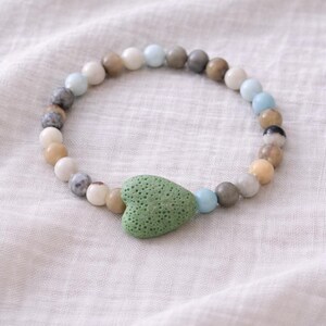 Puede incluir: Un brazalete de cuentas con un dije en forma de corazón verde. El brazalete está hecho de cuentas de varios colores, incluyendo blanco, beige, gris y azul. Se muestra sobre una tela blanca texturizada.
