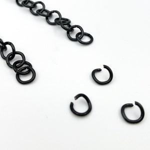 May include: A close-up of four black jump rings. The jump rings are small and round, and they are arranged in a scattered pattern on a white surface.
