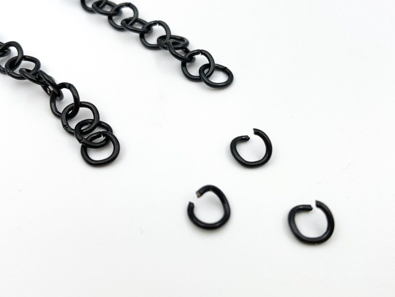 May include: A close-up of four black jump rings. The jump rings are small and round, and they are arranged in a scattered pattern on a white surface.