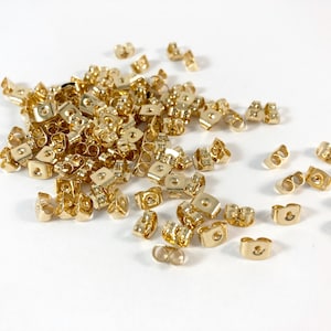 May include: A large pile of gold-colored earring backs. The backs are made of metal and have a small loop on the top for attaching earrings.