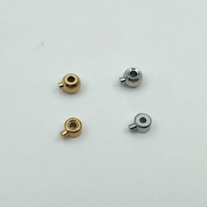 May include: Four small, metallic beads with a rounded shape and a small, protruding stem. Two beads are gold-colored, and two are silver-colored. Each bead has a small hole. The beads are arranged on a white surface.