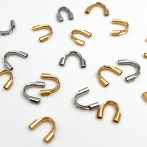 May include: A collection of gold and silver metal crimp beads. The beads are shaped like a U and have a small opening at the top. They are used for jewelry making.