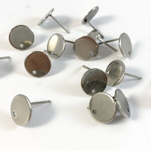 May include: A close-up of a group of silver earring blanks. The blanks are round with a small hole in the center. They are scattered on a white surface.