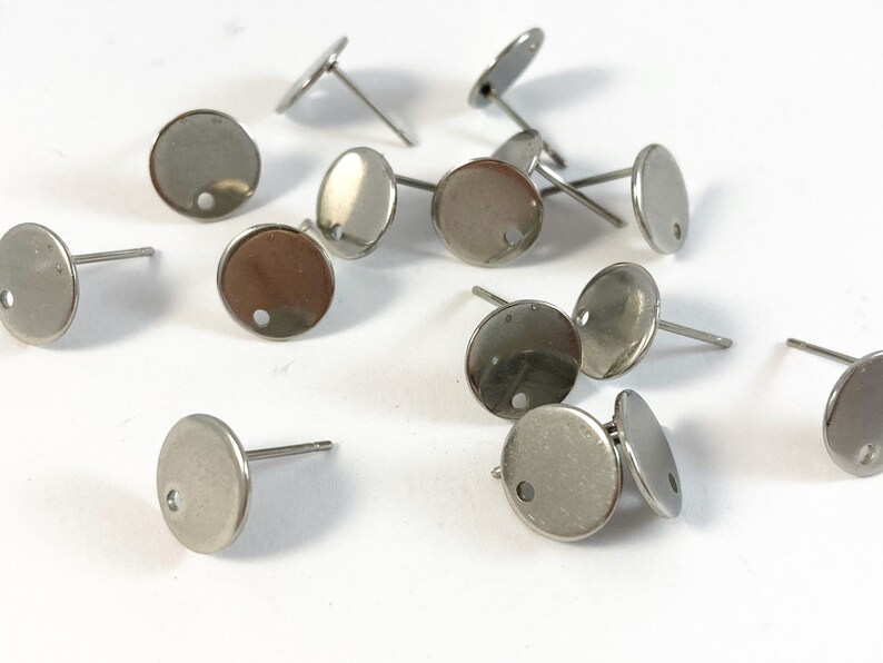 May include: A close-up of a group of silver earring blanks. The blanks are round with a small hole in the center. They are scattered on a white surface.