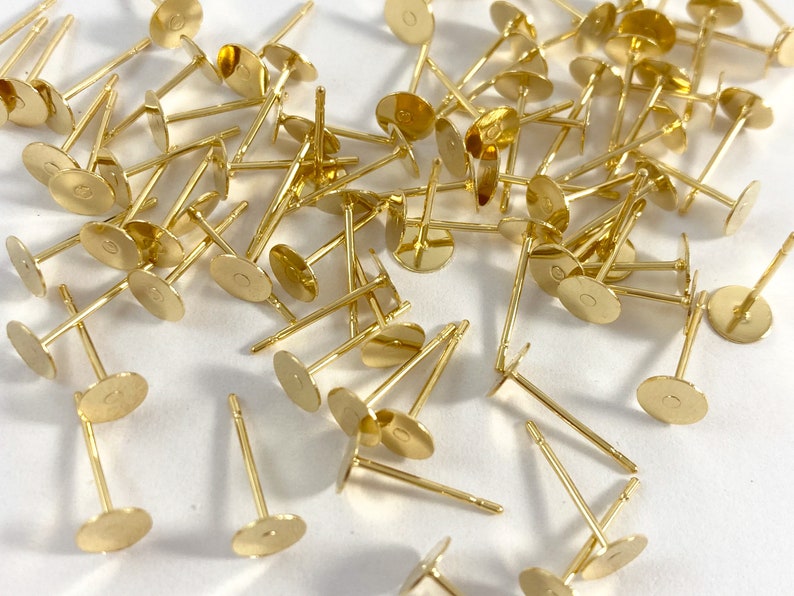 May include: A large quantity of gold-toned earring posts with flat round backs. The posts are scattered on a white surface.
