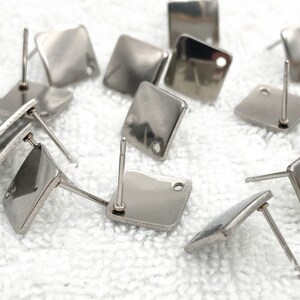 May include: A close-up of a group of silver square earring posts. The posts are arranged in a random pattern on a white surface.