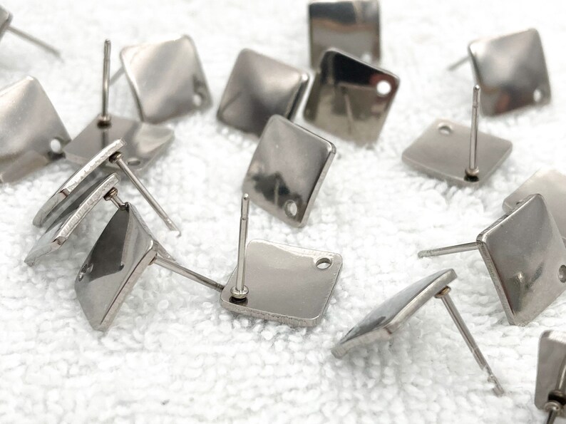 May include: A close-up of a group of silver square earring posts. The posts are arranged in a random pattern on a white surface.