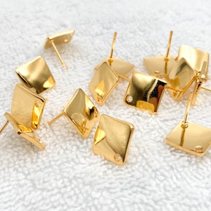 May include: A group of gold-toned square earring blanks with a small hole in the centre of each square. The earring blanks are scattered on a white surface.